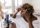 beauty trends and tips - A woman applying makeup while recording her beauty routine on smartphone indoors.