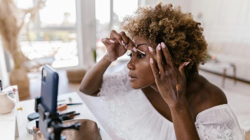 beauty trends and tips - A woman applying makeup while recording her beauty routine on smartphone indoors.