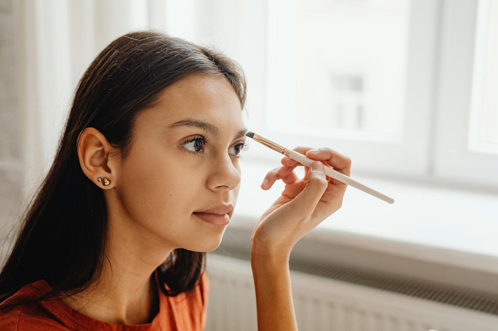beauty trends and tips - A young woman skillfully applying makeup with a brush by a window indoors.