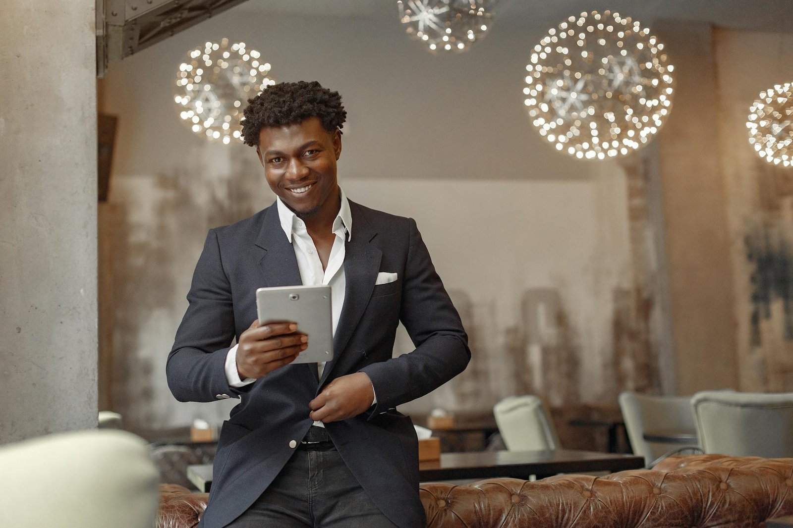 best gadgets for entrepreneurs - Positive successful black businessman in elegant formal outfit leaning on leather sofa and browsing tablet while unbuttoning jacket button in modern cafe
