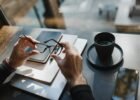 best gadgets for entrepreneurs - From above of crop unrecognizable man holding eyeglasses and sitting at wooden table with notebooks and cup of drink