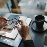 best gadgets for entrepreneurs - From above of crop unrecognizable man holding eyeglasses and sitting at wooden table with notebooks and cup of drink