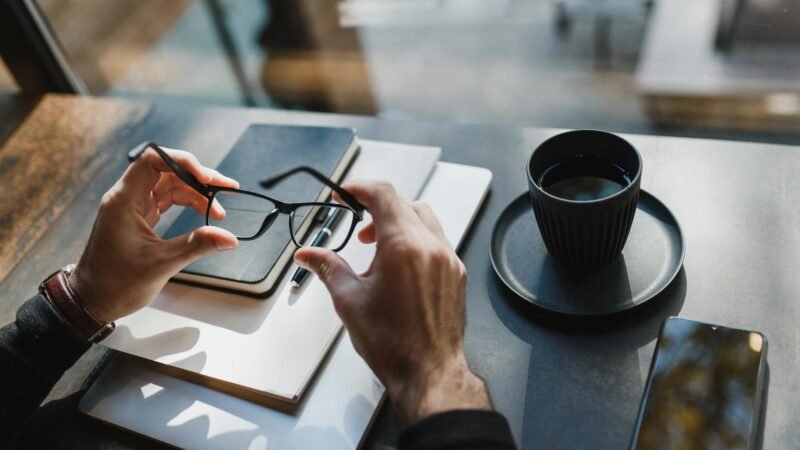 best gadgets for entrepreneurs - From above of crop unrecognizable man holding eyeglasses and sitting at wooden table with notebooks and cup of drink