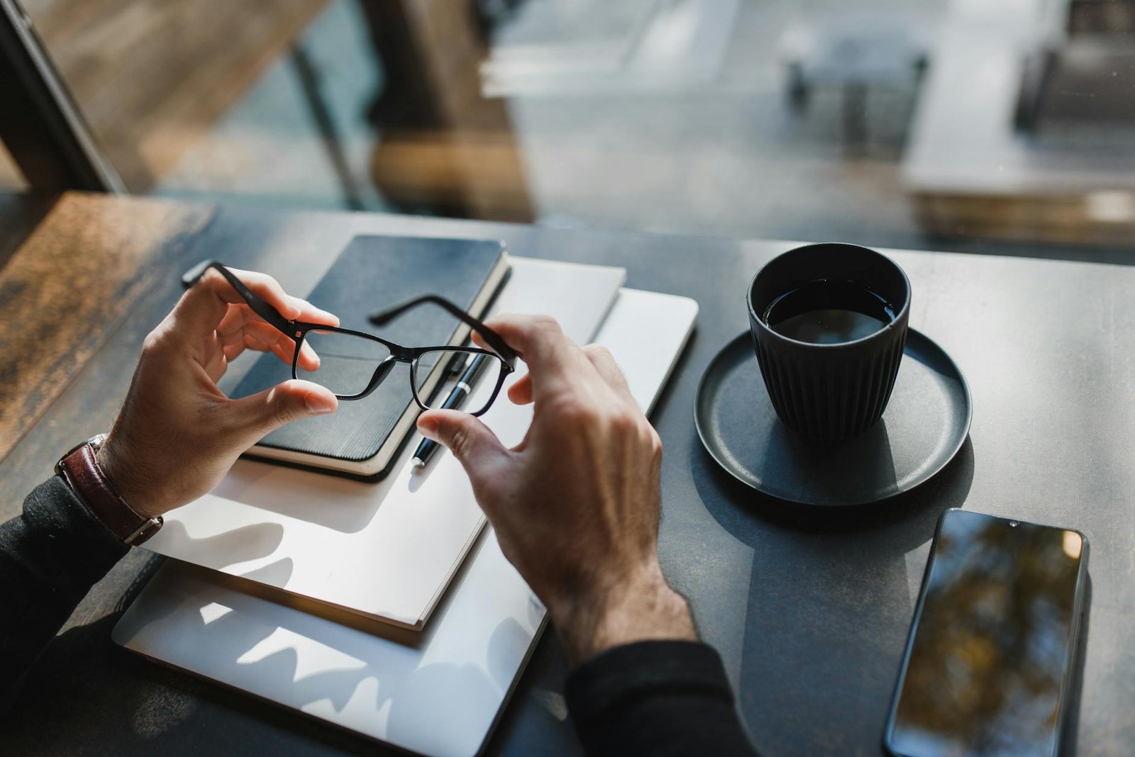 best gadgets for entrepreneurs - From above of crop unrecognizable man holding eyeglasses and sitting at wooden table with notebooks and cup of drink