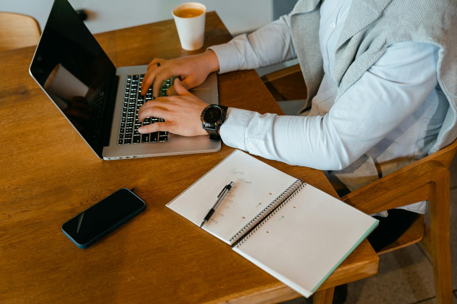 best gadgets for entrepreneurs - Person using laptop with notebook and coffee on a wooden table.