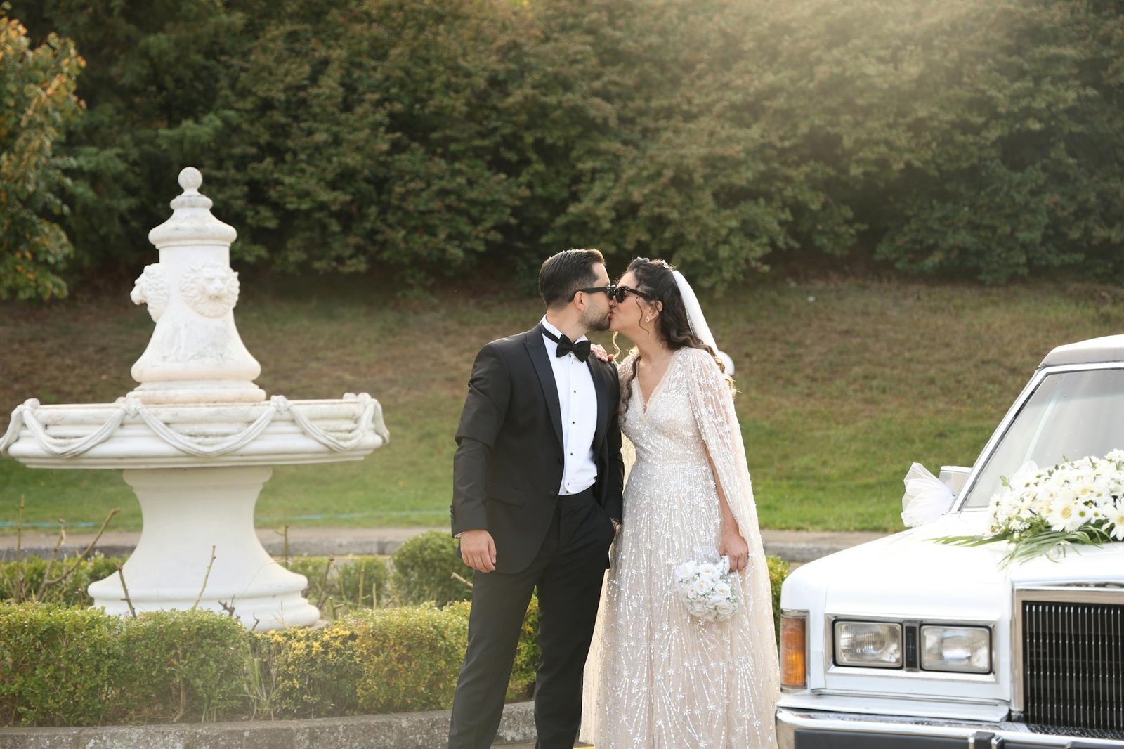 celebrity fashion moments - Bride and groom share a romantic kiss outdoors beside a classic car and fountain.