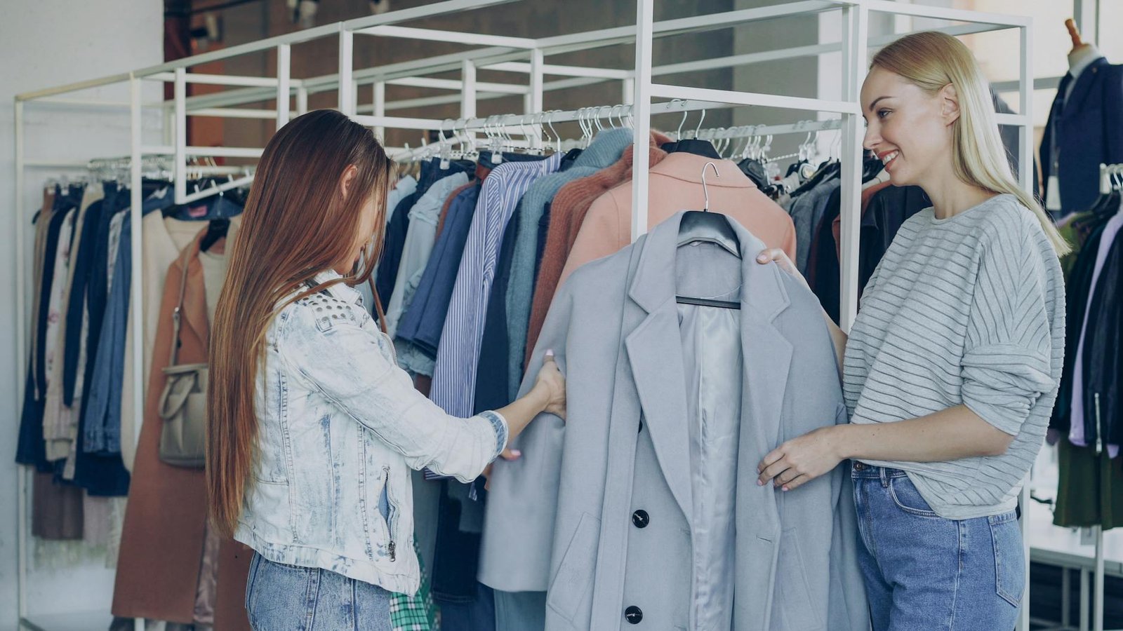 fashion trends for women - Two women shopping for jackets in a stylish clothing store, examining coats on a rack.