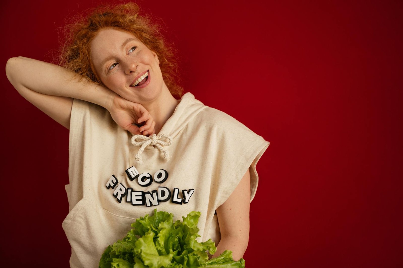 what is sustainable fashion - Cheerful woman with red hair in eco-friendly attire holding fresh lettuce against a vibrant red background.